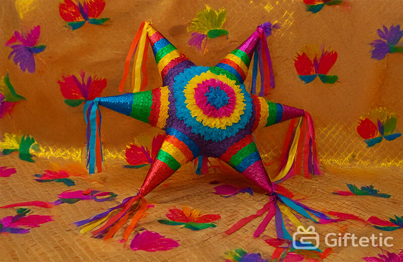 A brightly colored seven-point star-shaped piñata rests on a woven mat, surrounded by festive paper flower cutouts. Each cone of the piñata is decorated in vibrant rainbow colors with long multicolored streamers, representing the traditional Mexican Las Posadas celebration. The backdrop features more colorful decorations, creating a joyful and cultural atmosphere.
